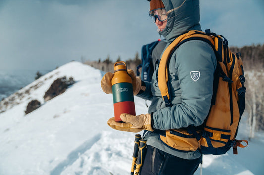 A bundled-up hiker holding an Adventure Flask beside a snowy mountain trail.